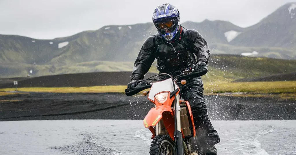 Piloto de motocicleta atravessando um trecho alagado durante chuva forte, usando equipamentos de proteção para pilotagem.