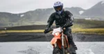 Piloto de motocicleta atravessando um trecho alagado durante chuva forte, usando equipamentos de proteção para pilotagem.